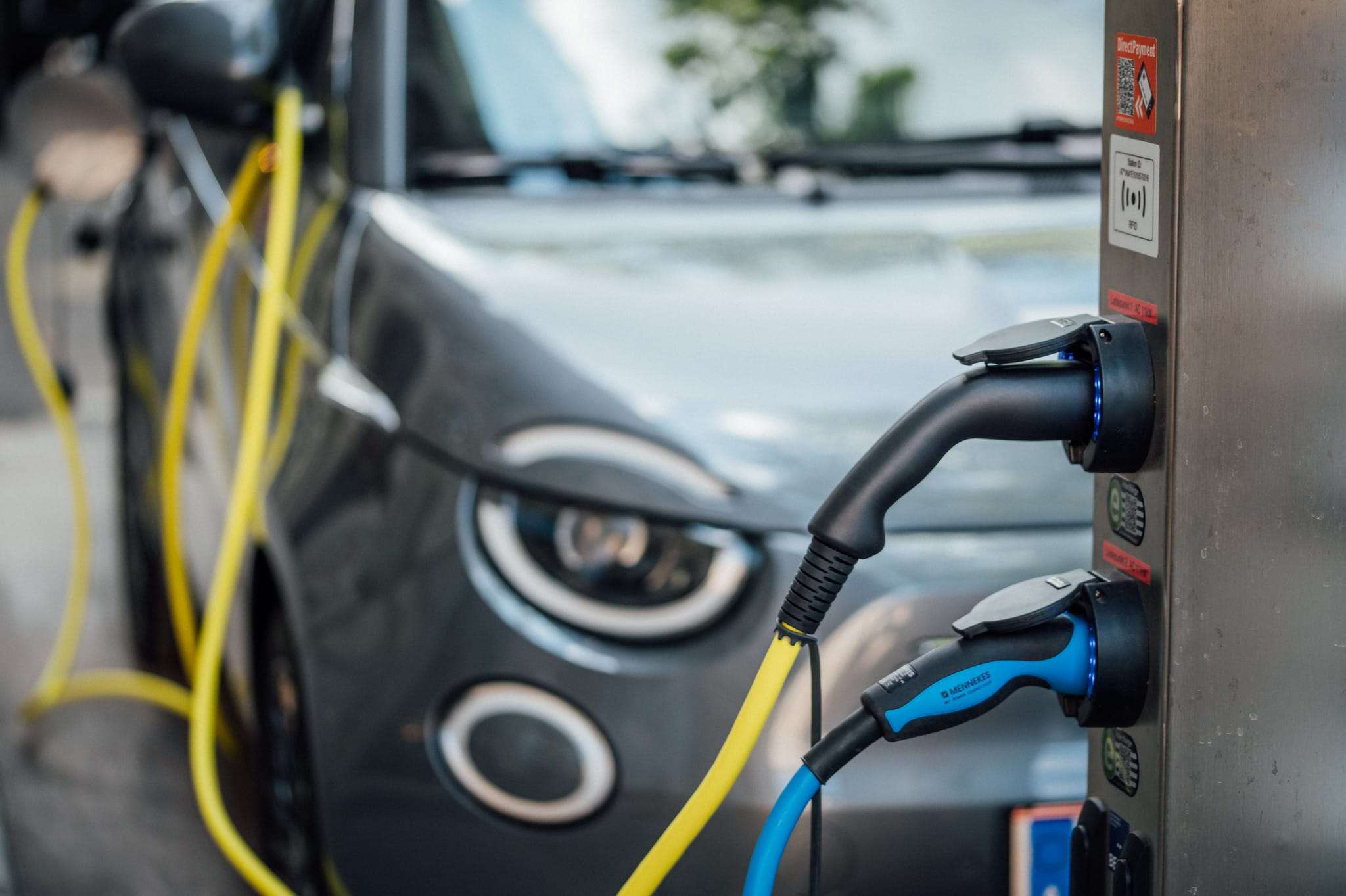 Power cables plugged into an electric vehicle charging station with an electric car in the blurry background - Image by Flickr user Ivan Radic