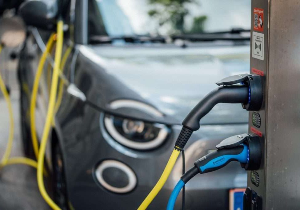 Power cables plugged into an electric vehicle charging station with an electric car in the blurry background - Image by Flickr user Ivan Radic
